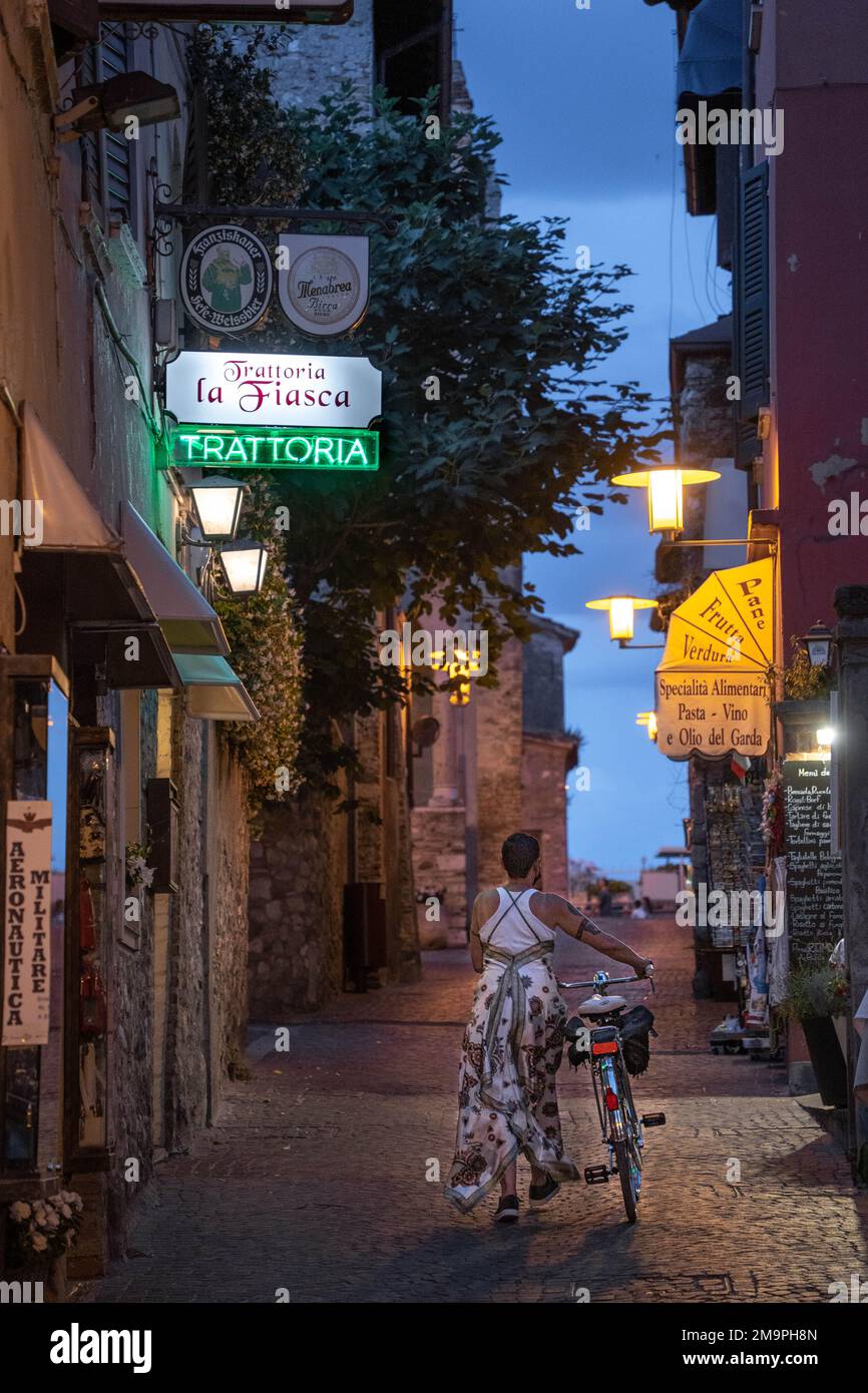 A woman walks with a bicycle down an empty narrow street illuminated by restaurant signs in Sermione, Italy Stock Photo