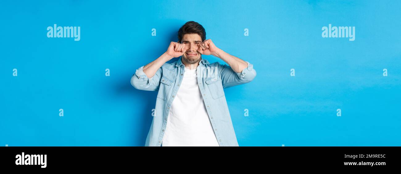 Sad timid man crying, wiping tears off face and sobbing, looking offended and upset, standing over blue background Stock Photo