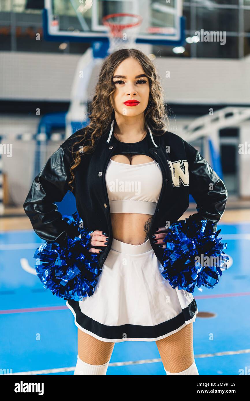 Brunette cheerleader in black and white uniform with a jacket posing with blue shiny pom-poms. Basketball court blurred in the background. High quality photo Stock Photo