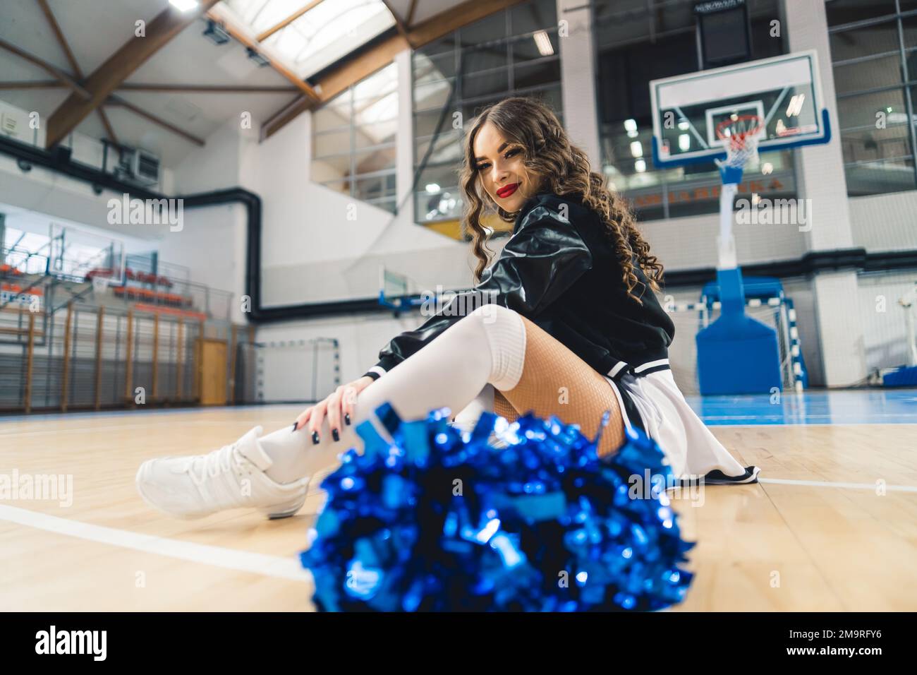 Brunette, curly-haired cheerleader in black and white uniform and jacket sitting on basketball court. Blue shiny pom-pom blurred in the foreground. High quality photo Stock Photo
