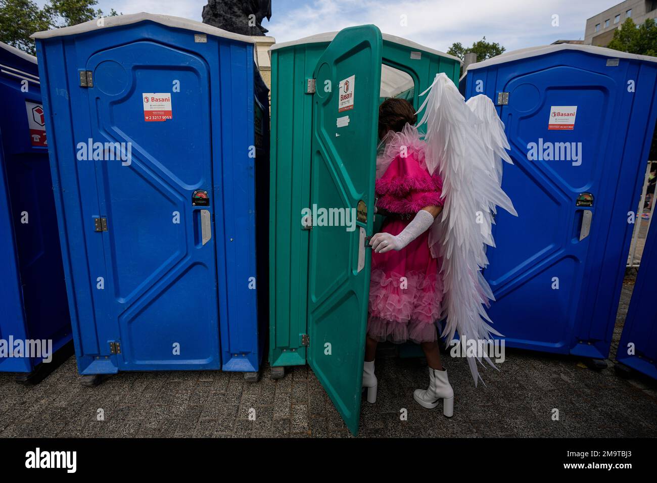 Gerson, wearing angel wings, enters a portable toilet set up for