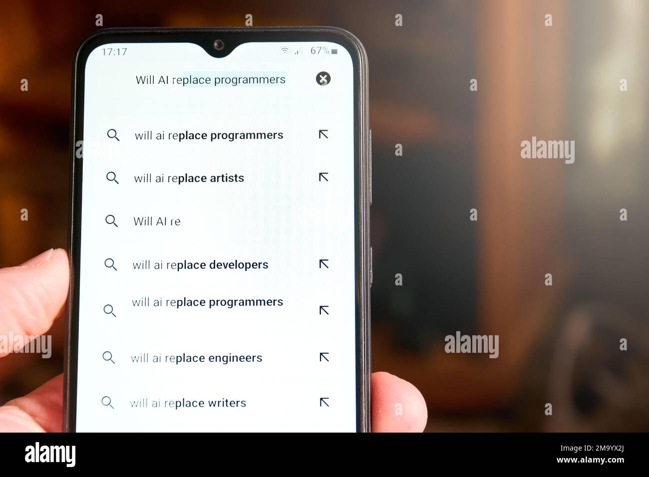 Horizontal close up of mobile phone screen with search 'Will AI replace programmers' and list of suggested searches below. People searches on AI, conc Stock Photo