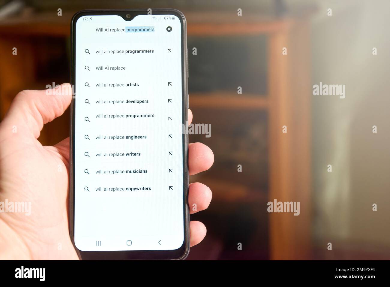 Horizontal shot of male hand holding mobile phone screen with search 'Will AI replace programmers' and list of suggested searches below. People search Stock Photo