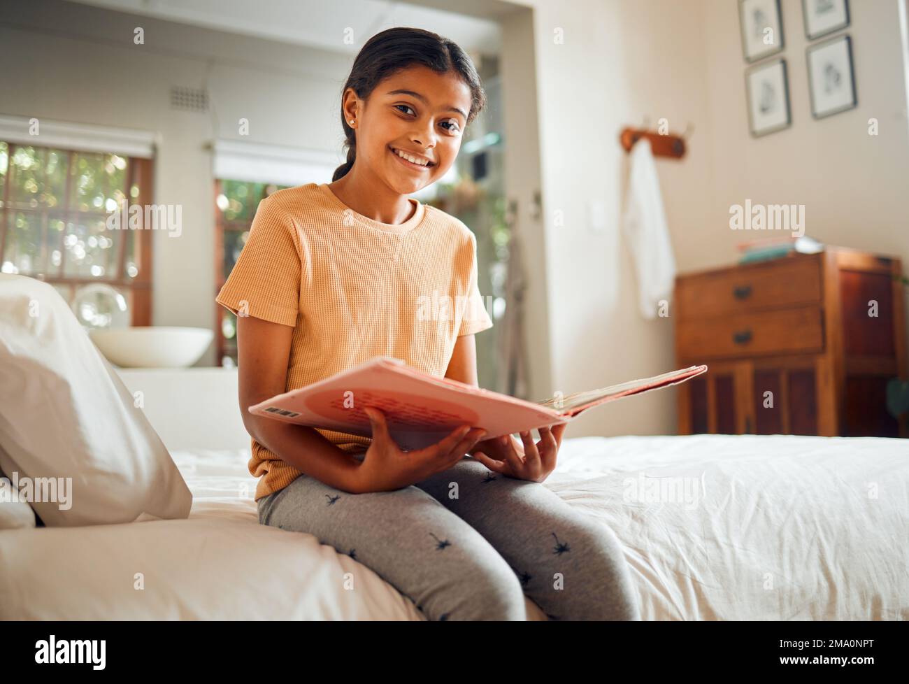 Happy little girl, reading book and bed with smile for story time, education or learning in comfort at home. Portrait of cute female child smiling in Stock Photo