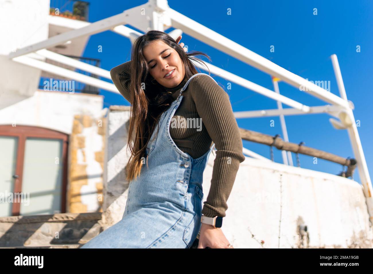 low-angle-content-ethnic-woman-in-denim-overall-touching-hair-with