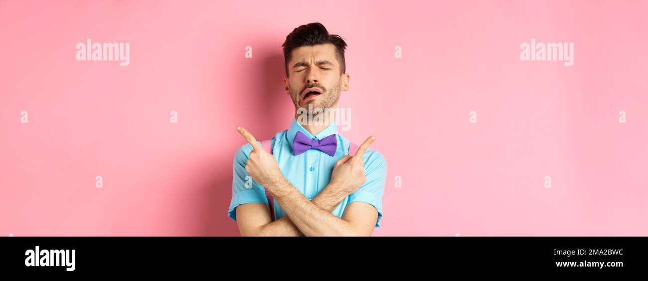 Sad and disappointed young man in bow-tie, crying and pointing fingers sideways, showing two things he wants, sobbing from unrequited love, standing Stock Photo
