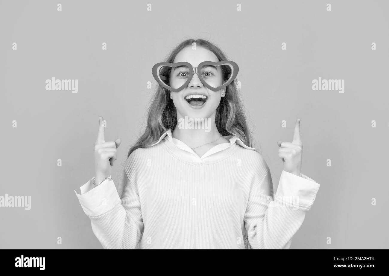 Happy girl in big funny heartshaped glasses pointing fingers up blue background, copy space