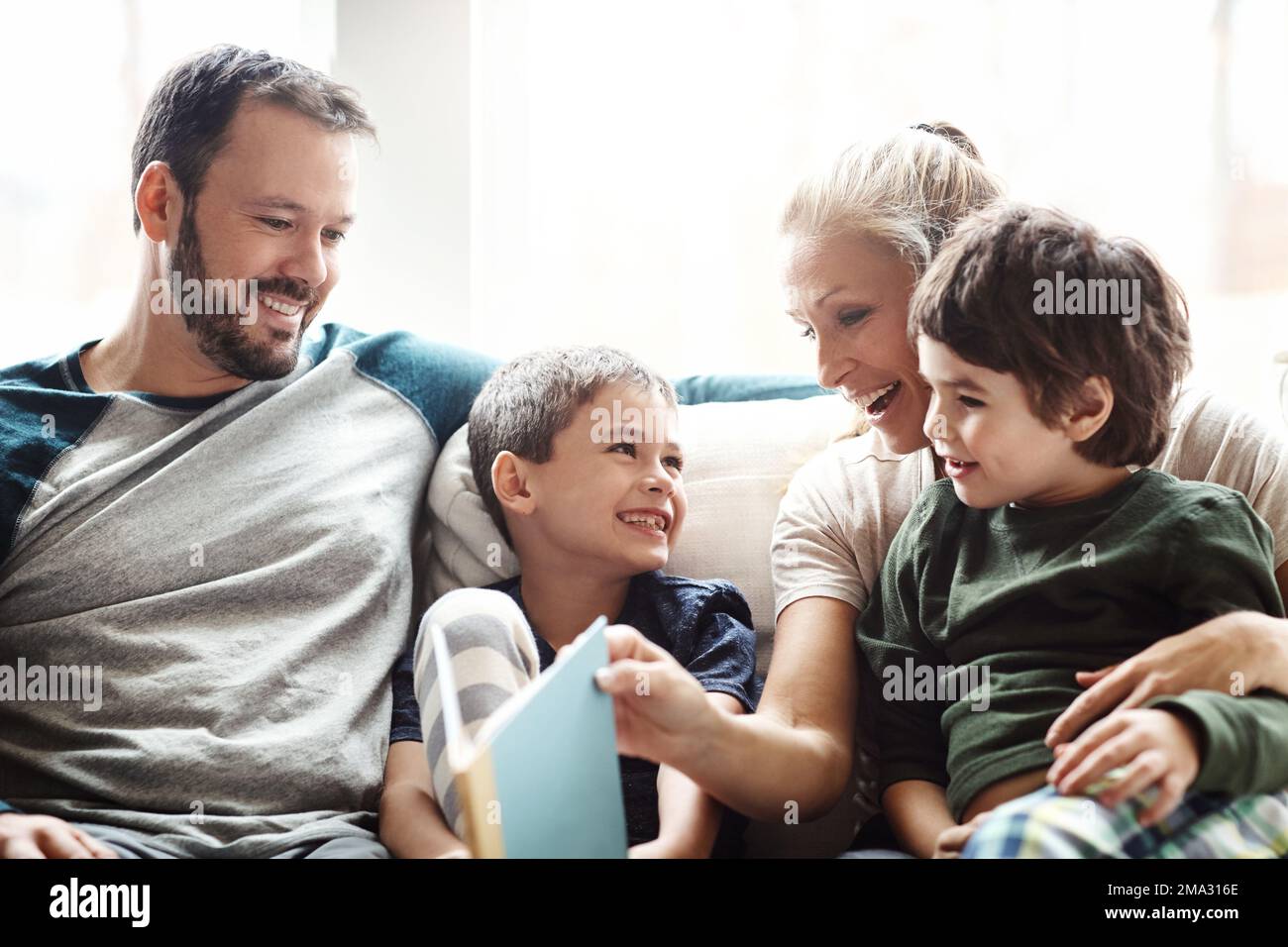Mother, father and kids on sofa for storytelling time in living room of happy family home. Love, books and couple with children smile, book and Stock Photo