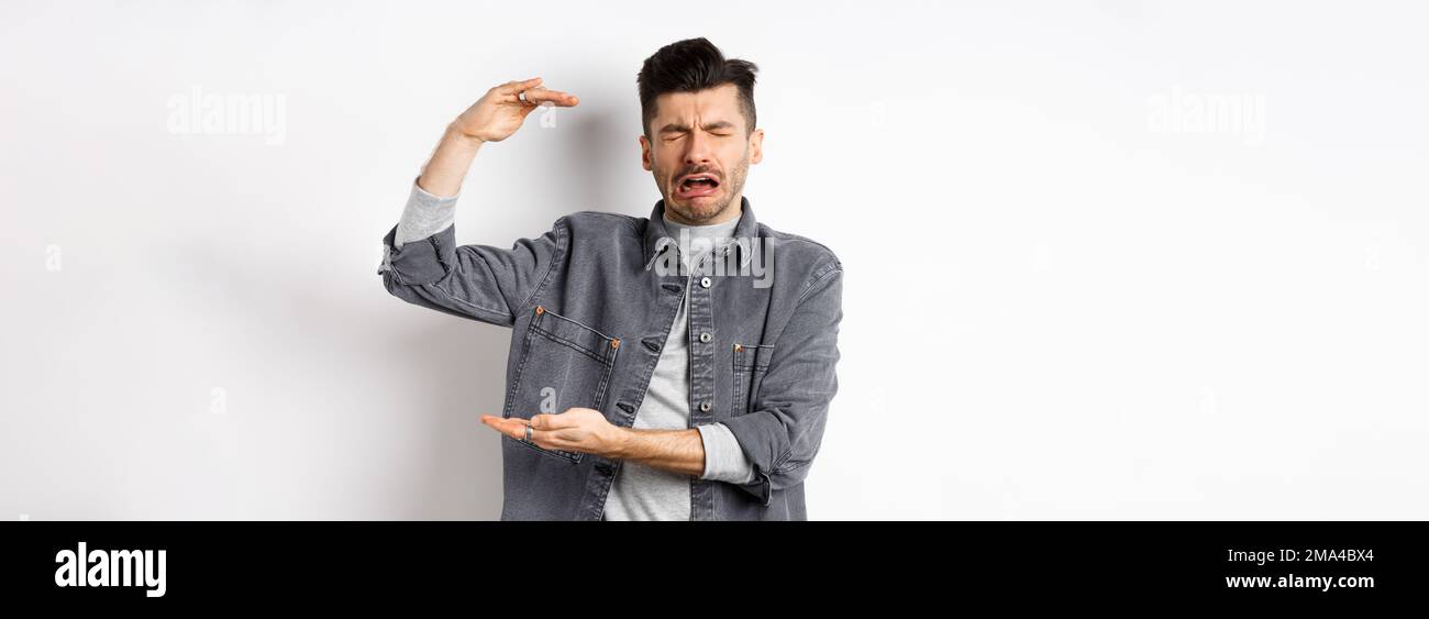 Sad man crying and showing big size object, shaping large thing and complaining, sobbing while standing on white background Stock Photo