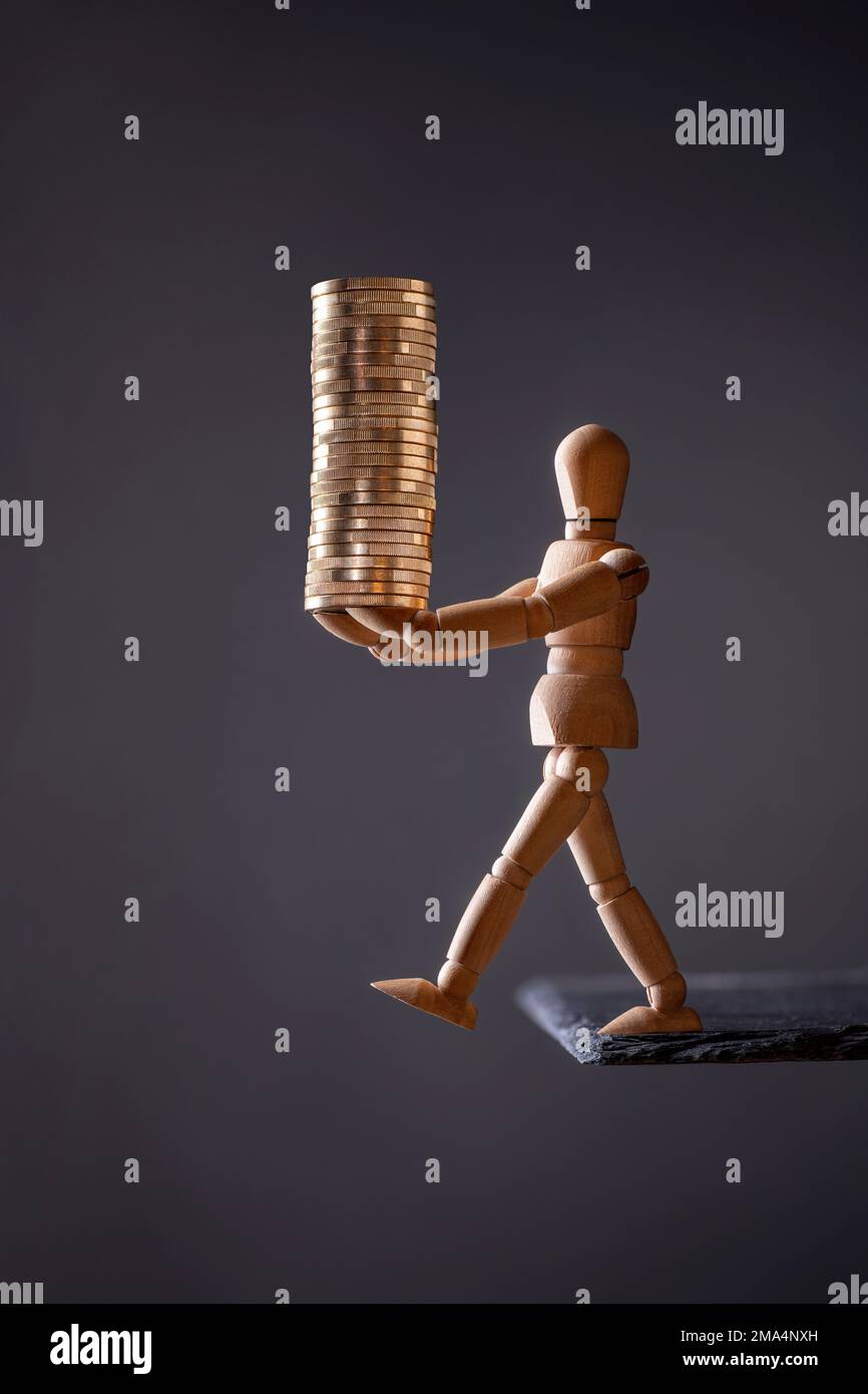 A wooden mannequin carries a stack of coins while threatening to fall off the edge of a stone slab Stock Photo