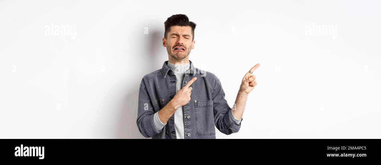 Sad crying man pointing fingers left at logo, sobbing and whining, being jealous or upset, standing on white background Stock Photo