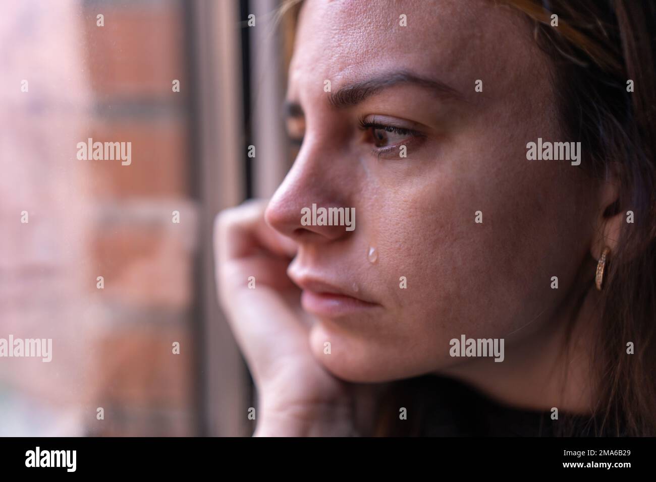 close-up of a depressed woman looking out the window, while a tear falls down her cheek. Depression and mental health concept Stock Photo