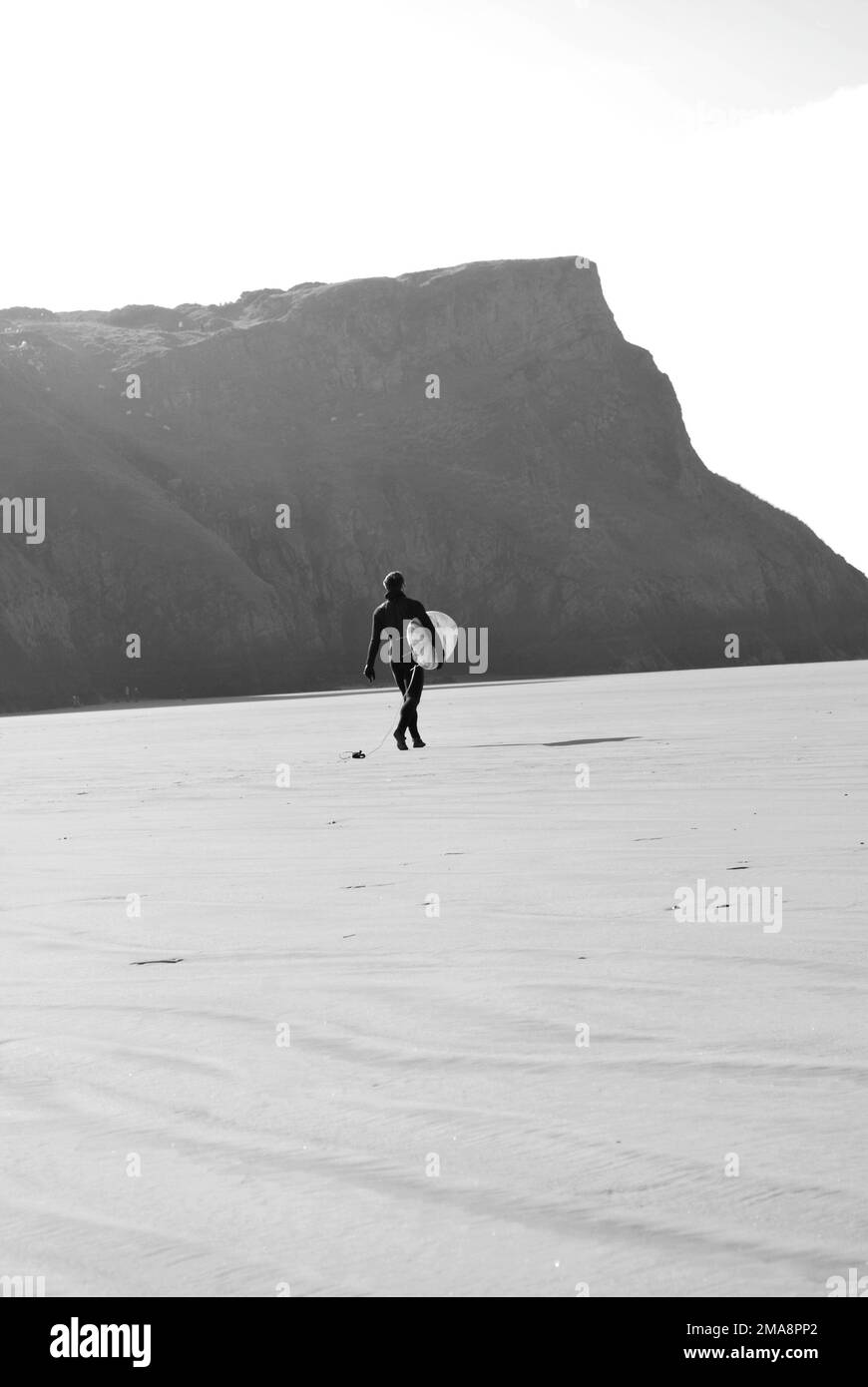 A vertical monochrome shot of a surfer walking along a sandy coast with a board Stock Photo