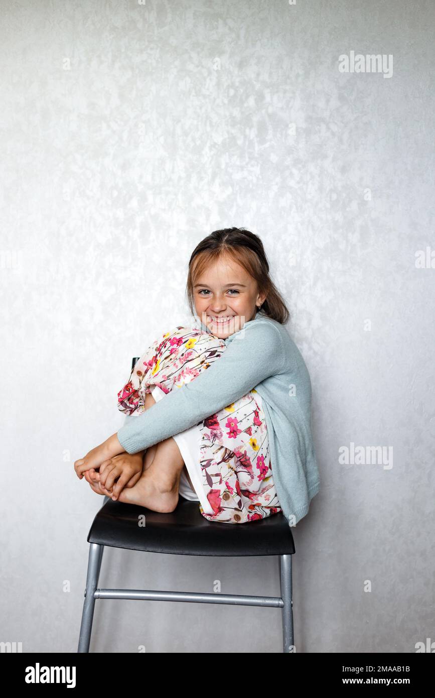 A happy smiling little girl is sitting on a stool with her legs pulled ...