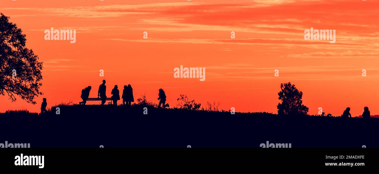 Group of young people enjoying beautiful summer sunset on a hill, silhouette of back lit male and female teenagers with orange sky in background Stock Photo
