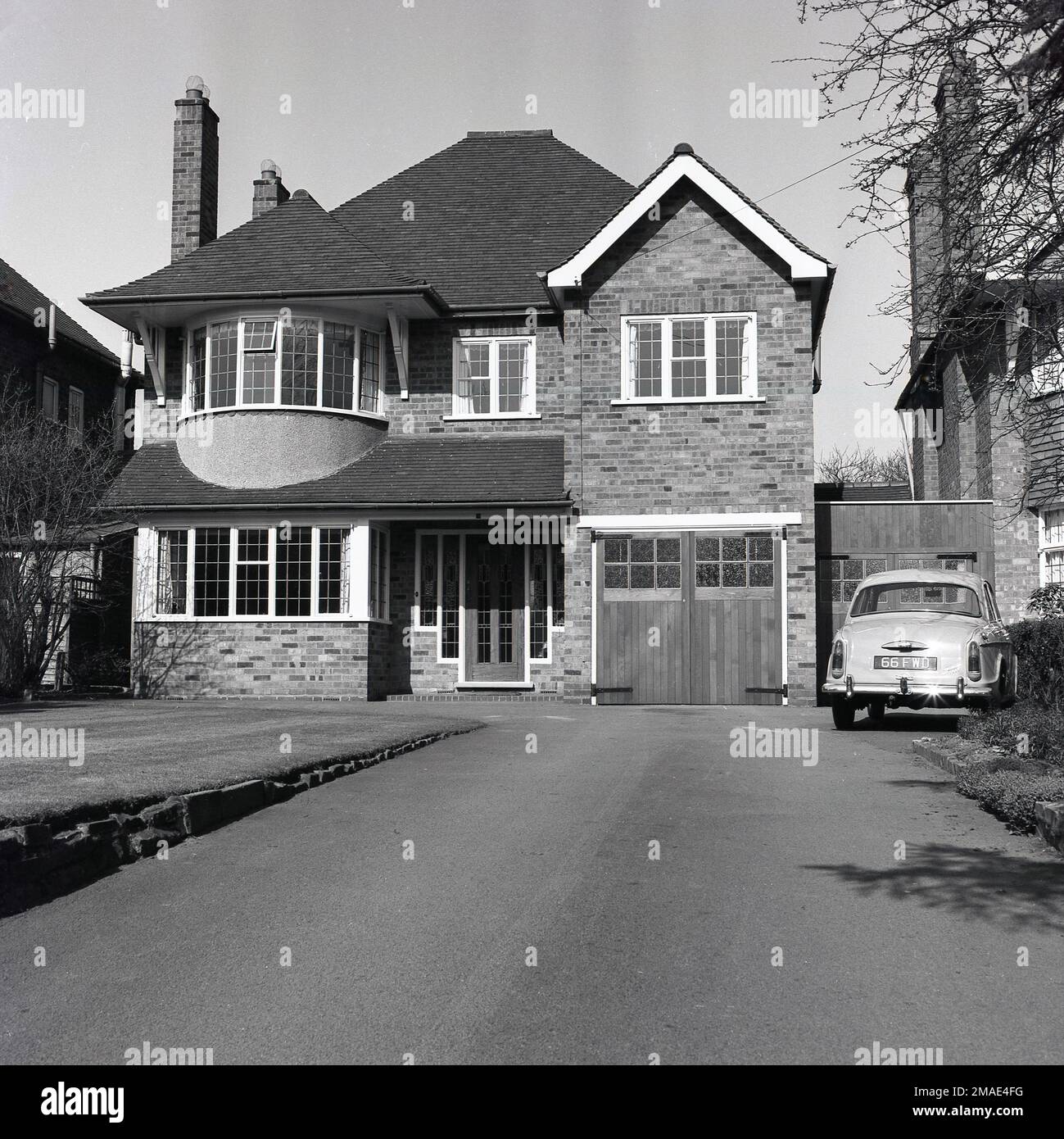 1960s, historical, front exterior view of a large suburban house, with internal garage, with wooden doors, England, UK.  A Woseley car of the era is parked in the driveway infront of second garage built on the side of the house and which is attached to the neighbouring property. Stock Photo