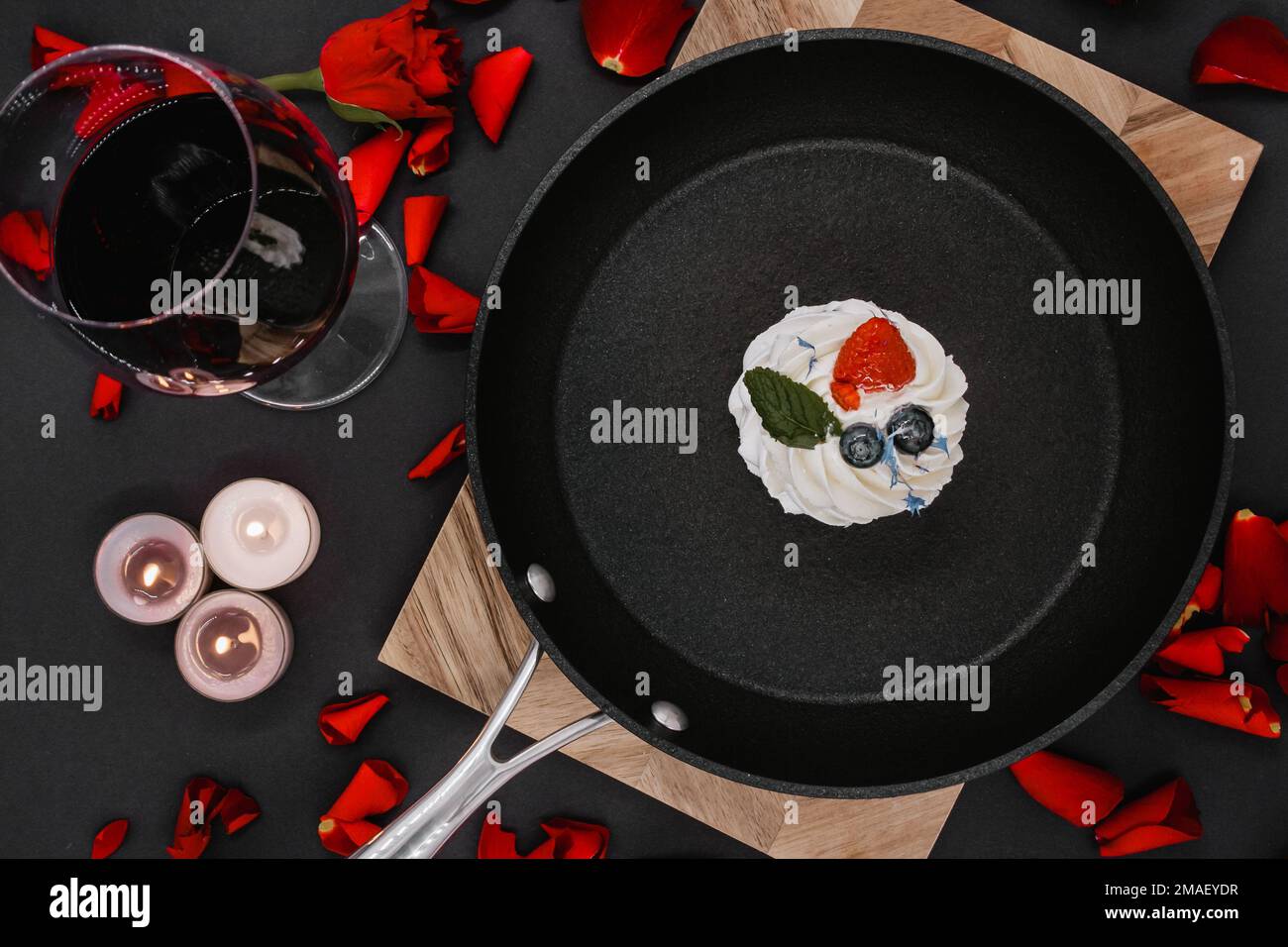 black frying pan with a pavlova cake on a wooden board with a black background, next to which is a glass of red wine and lit candles. flat lay Stock Photo