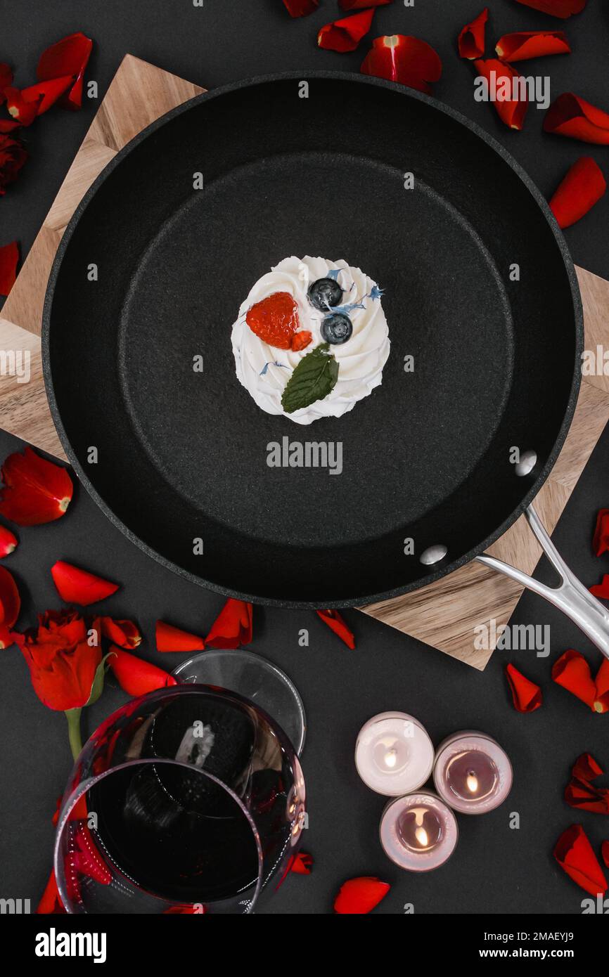 black frying pan with a pavlova cake and scattered red rose petals on a wooden board with a black background, next to which is a glass of red wine and Stock Photo