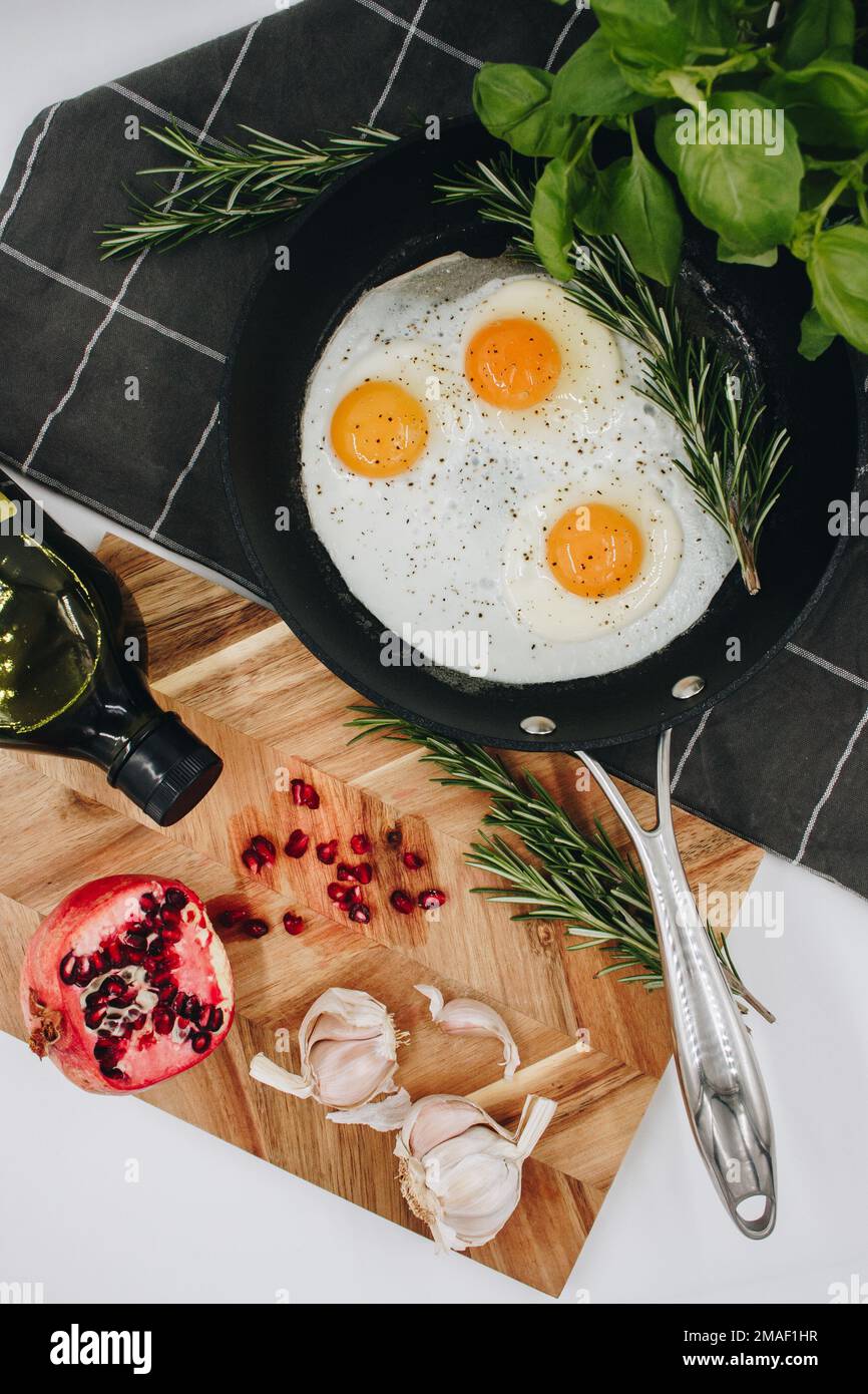 black frying pan on a black towel with three fried egg yolks in it next to a wooden cutting board with a red pomegranate on it. Stock Photo