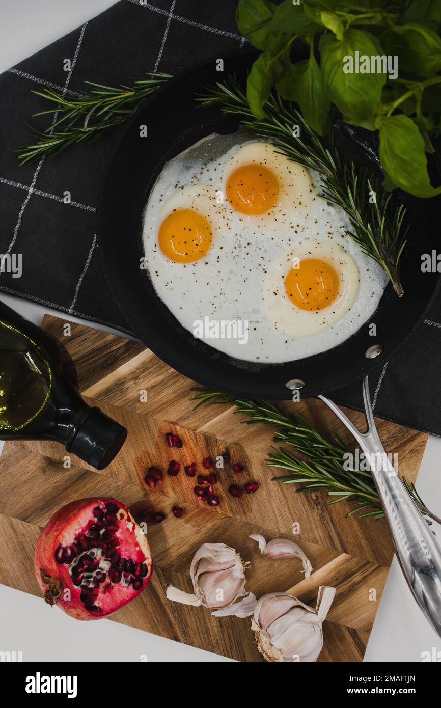 black frying pan on a black towel with three fried egg yolks in it next to a wooden cutting board with a red pomegranate on it. Stock Photo