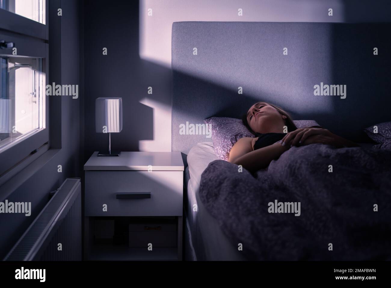 Sleeping woman in bed at night in dark bedroom. Person under blanket and cover. Person asleep, resting in cold room at home. Tired lady. Stock Photo