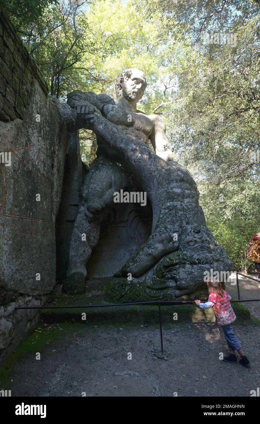 A kid near a huge statue depicting Hercules fighting Cacus also called Sacred Grove in the Park of the Monsters of Bomarzo. Viterbo,Italy Stock Photo