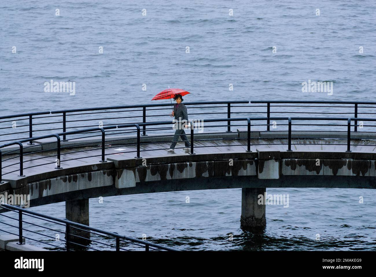An Asian woman walking across an ocean concrete pier or jetty carrying a red umbrella Stock Photo