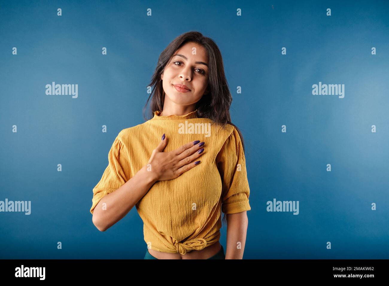 Portrait of young brown-haired woman wearing yellow top isolated over blue background touching chest with her palm, smiles tenderly and looks at the c Stock Photo