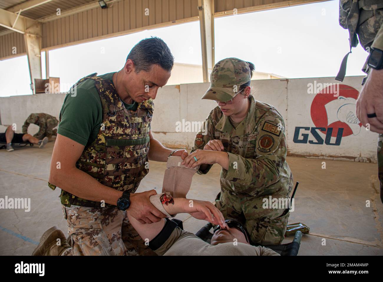 U.S. Air Force Staff Sgt. Elissa Gray, right, paramedic, 386th Expeditionary Medical Group, explains to an Italian Army coalition partner, how to properly apply a bandage to control simulated massive bleeding on Staff Sgt. Matthew Depodesta, firefighter, 386th Expeditionary Civil Engineer Squadron, during the joint coalition Tactical Combat Casualty Care course at a skating rink at Ali Al Salem Air Base, Kuwait, May 29, 2022. In the TCCC course, which replaced the Self-Aid Buddy Care course, U.S. service members and coalition partners practiced applying a tourniquet, wrapping a bandage and ope Stock Photo