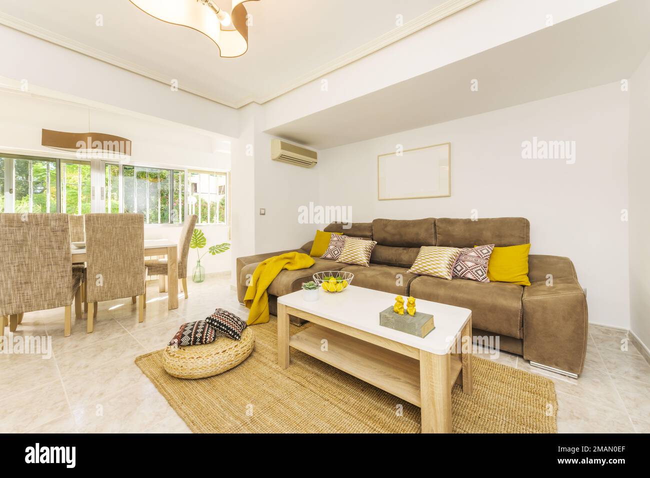 Living room furnished with light, brown and yellow tones, with a brown leatherette sofa, yellow blankets and cushions and a window next to the dining Stock Photo