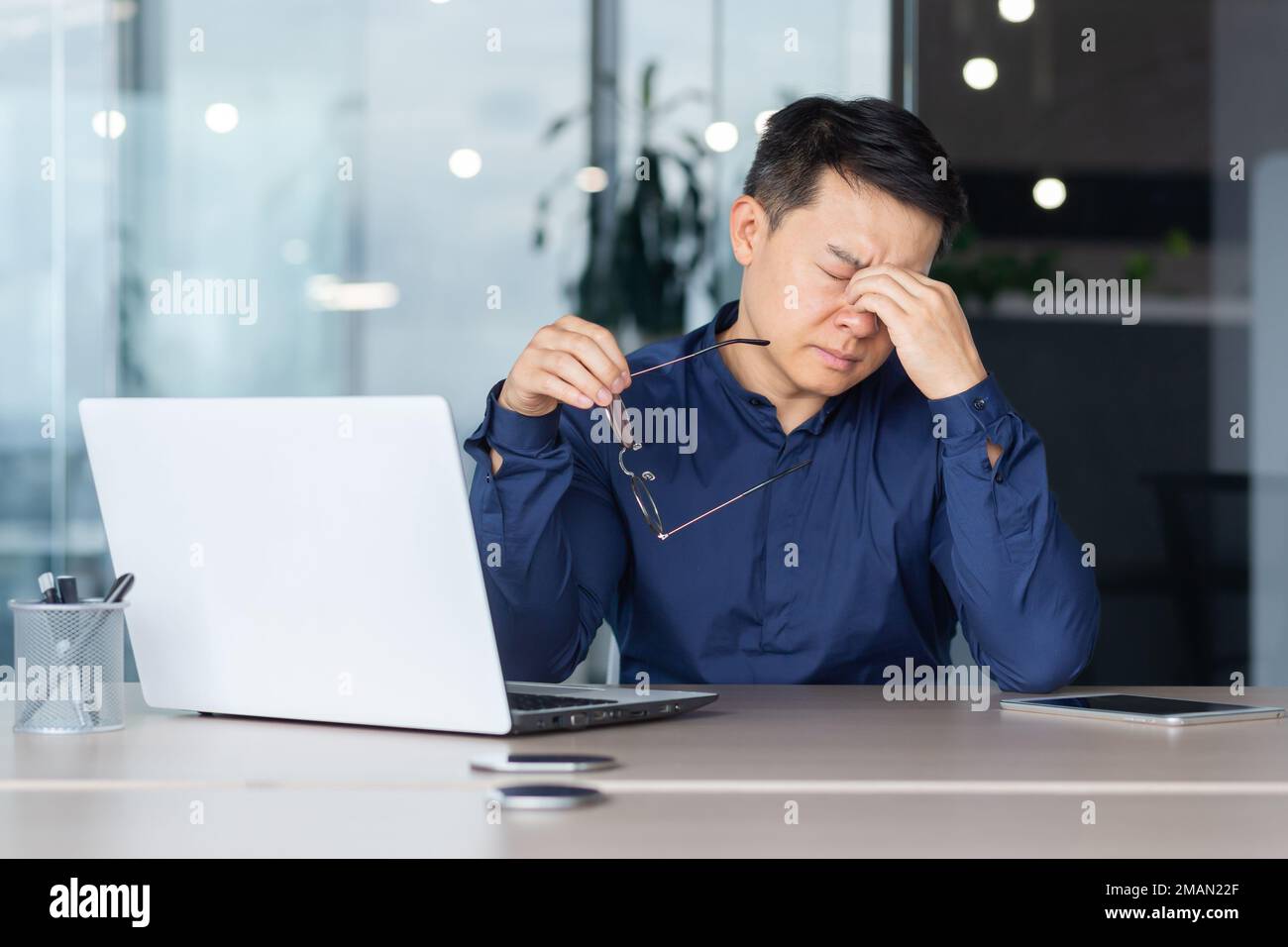 Asian worker tired at work has pain in eyes, businessman in glasses working late, using laptop at work inside office, serious and concentrated pensive. Stock Photo