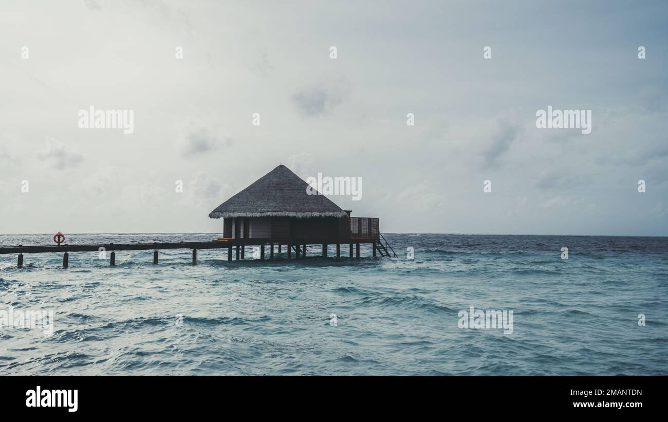 A view of an overwater bungalow with a pyramid hip roof, over the crystalline turquoise waters of the ocean water that lap gently at the deck of the l Stock Photo