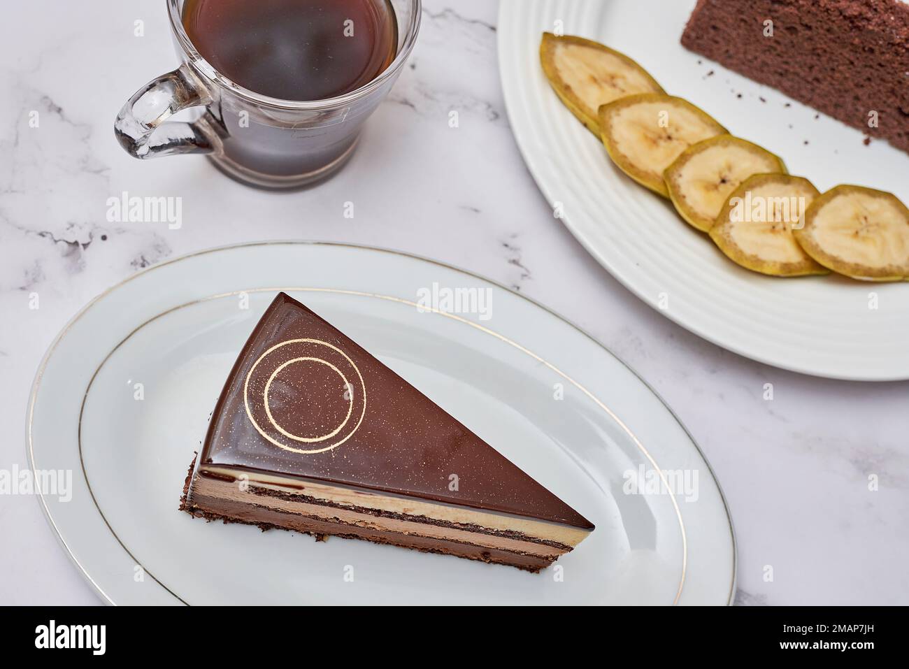 a piece of chocolate cake on a plate with a cup of coffee and banana slices in the photo is taken Stock Photo