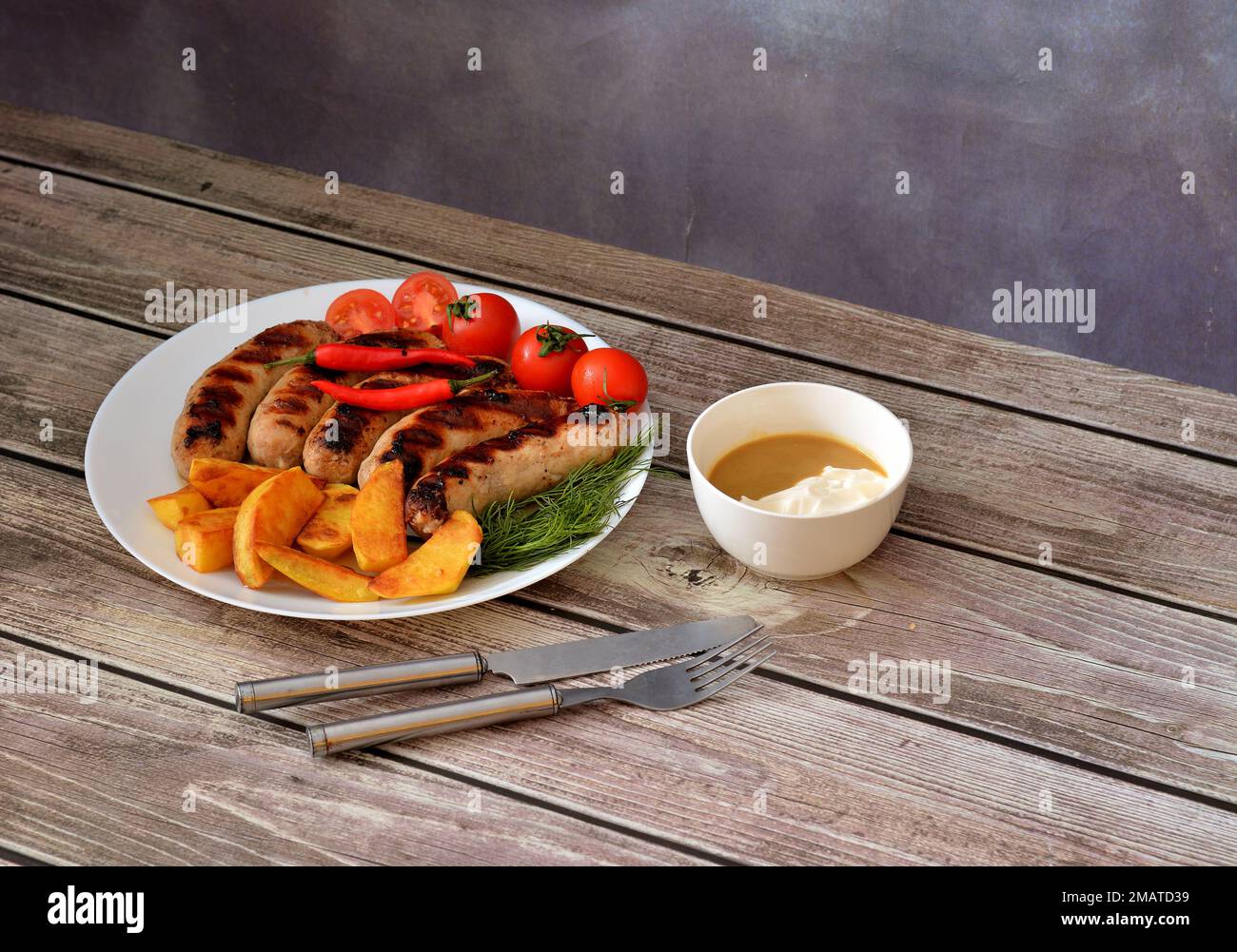 A plate with several grilled sausages, cherry tomatoes, hot peppers, fried potatoes and herbs on a wooden table, next to a cup with sauce and cutlery. Stock Photo