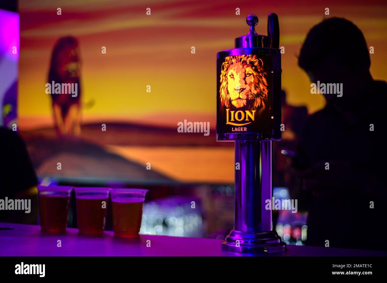 Weligama, Sri Lanka - 09 29 2022: Lion lager backlit logo on a liquor pub table and 3 beer cups. Silhouetted bartender in the background. Stock Photo