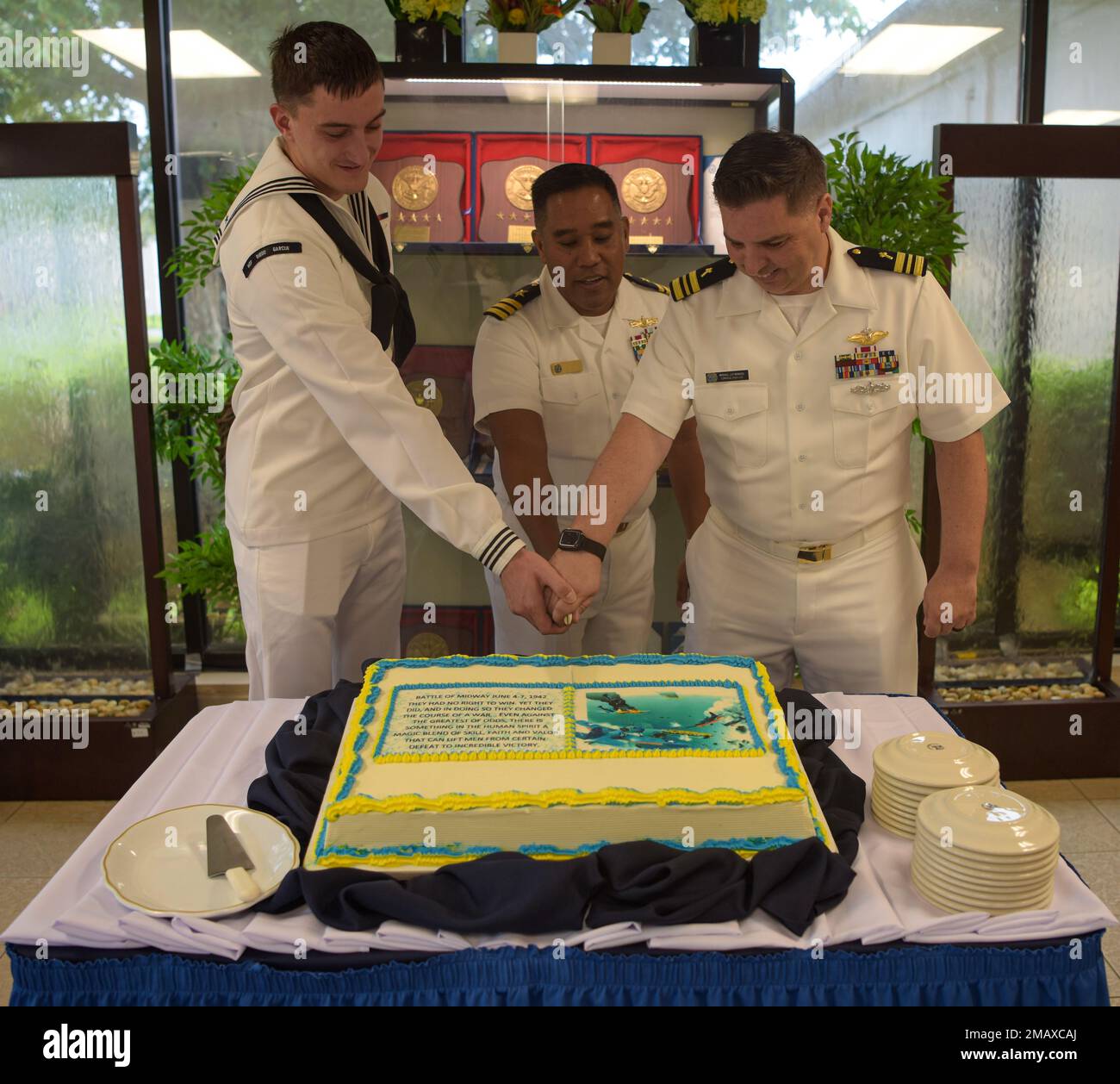220607-N-EJ241-1021    DIEGO GARCIA, British Indian Ocean Territory (June 7, 2022) – Brady A. Stuart, assigned to U.S. Navy Support Facility (NSF) Diego Garcia, left, Cmdr. Robert E. Bulatao, executive officer of NSF Diego Garcia, center, and NSF Diego Garcia Command Chaplain, Lt. Cmdr. Michael J. Monroig, cut a cake during an 80th anniversary commemoration of the Battle of Midway. Our remembrance of Midway illustrates how our battle achievements in innovation, intelligence, and courage serve as a model and as inspiration as we face the challenges of the future. Stock Photo