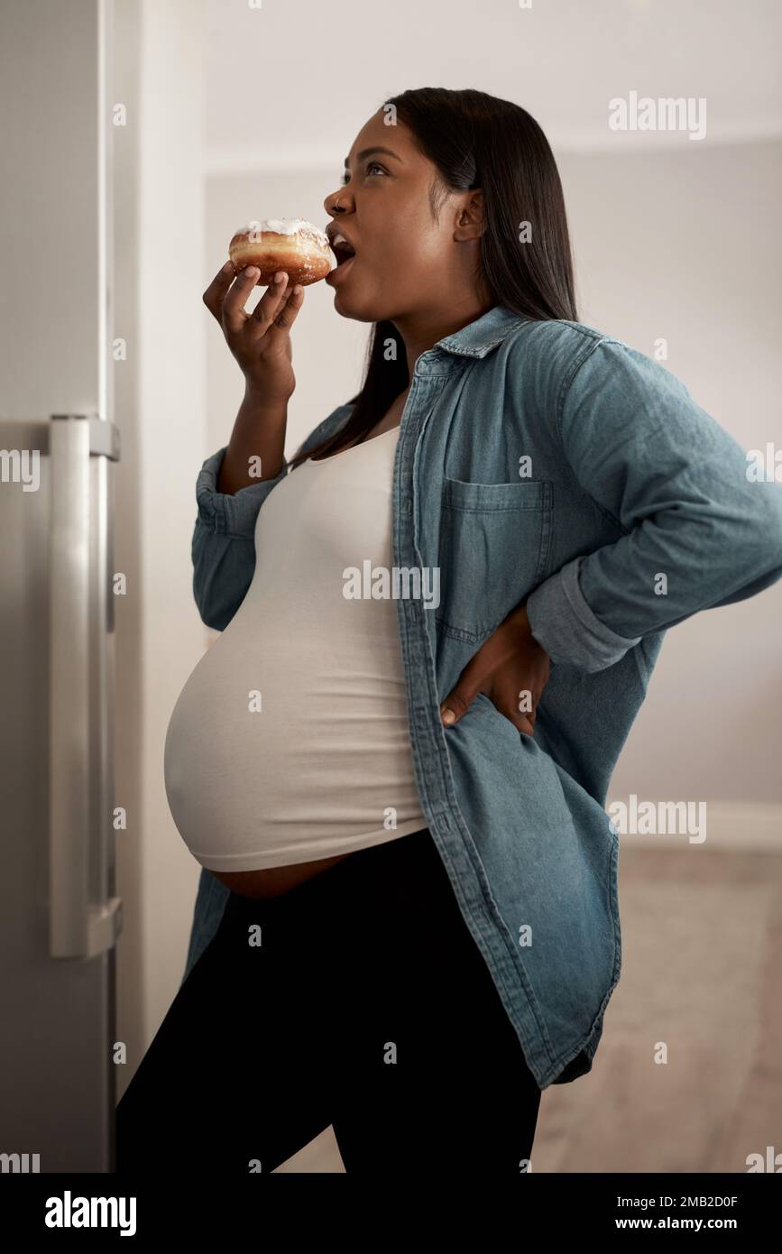 The cravings are kicking me hard today. a pregnant woman eating a donut at home. Stock Photo