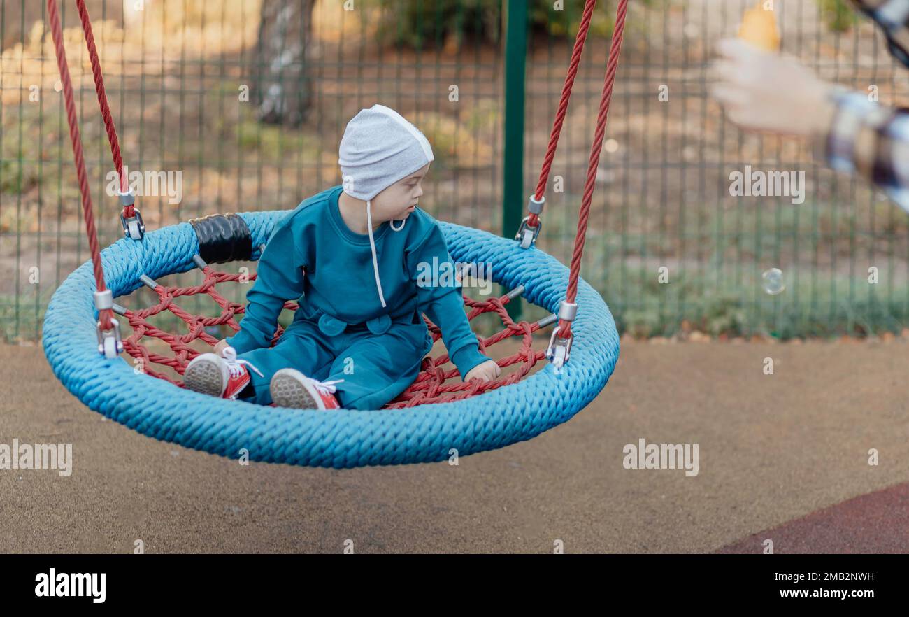 Cute Little boy with down syndrome in a funny hat walks in the playgroundk, swinging on a swing Stock Photo
