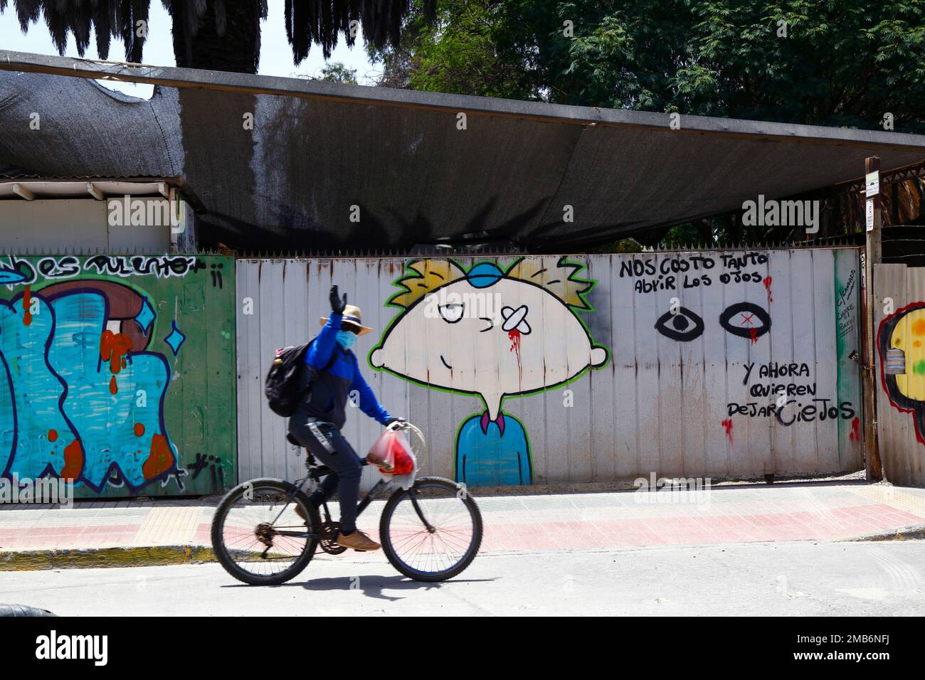 Man cycling past mural protesting against the number of eye injuries inflicted by riot police on protestors during the Chilean protests of 2019-2020, Copiapo, Region III, Chile. The Spanish translates as: 'it cost us so much to open our eyes, and now they want to leave us blind'. Stock Photo