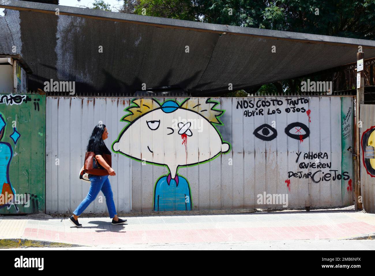 Woman walking past mural protesting against the number of eye injuries inflicted by riot police on protestors during the Chilean protests of 2019-2020, Copiapo, Region III, Chile. The Spanish translates as: 'it cost us so much to open our eyes, and now they want to leave us blind'. Stock Photo