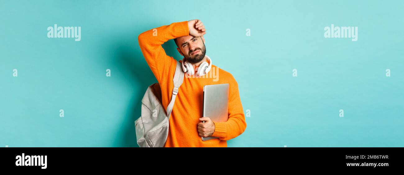 Tired male student wiping sweat off forehead, holding laptop and ...