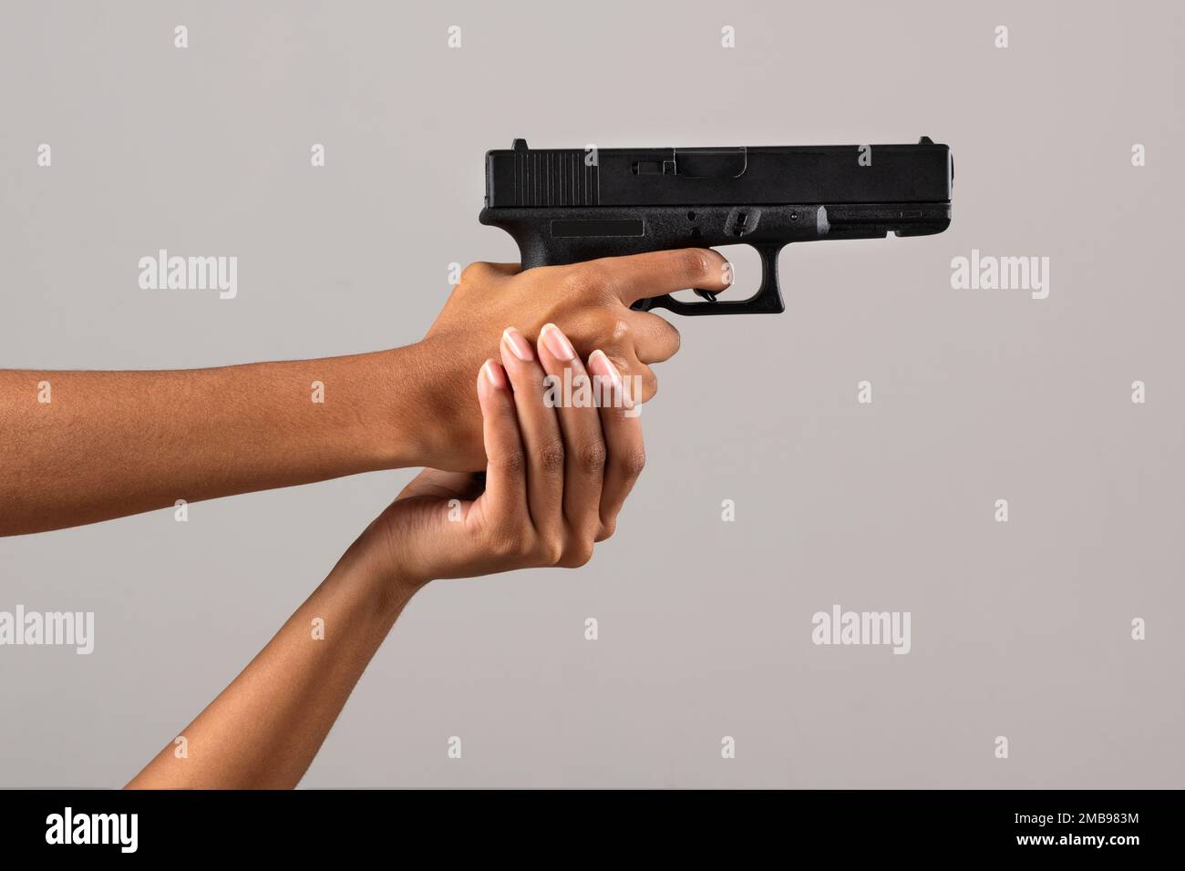 Crop anonymous African American woman with black gun pulling trigger on gray background Stock Photo
