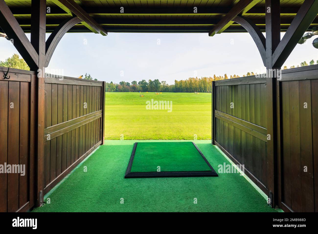 Entrance with wooden fence and roof to modern golf driving range with big green field and trees under cloudy sky on sunny day Stock Photo