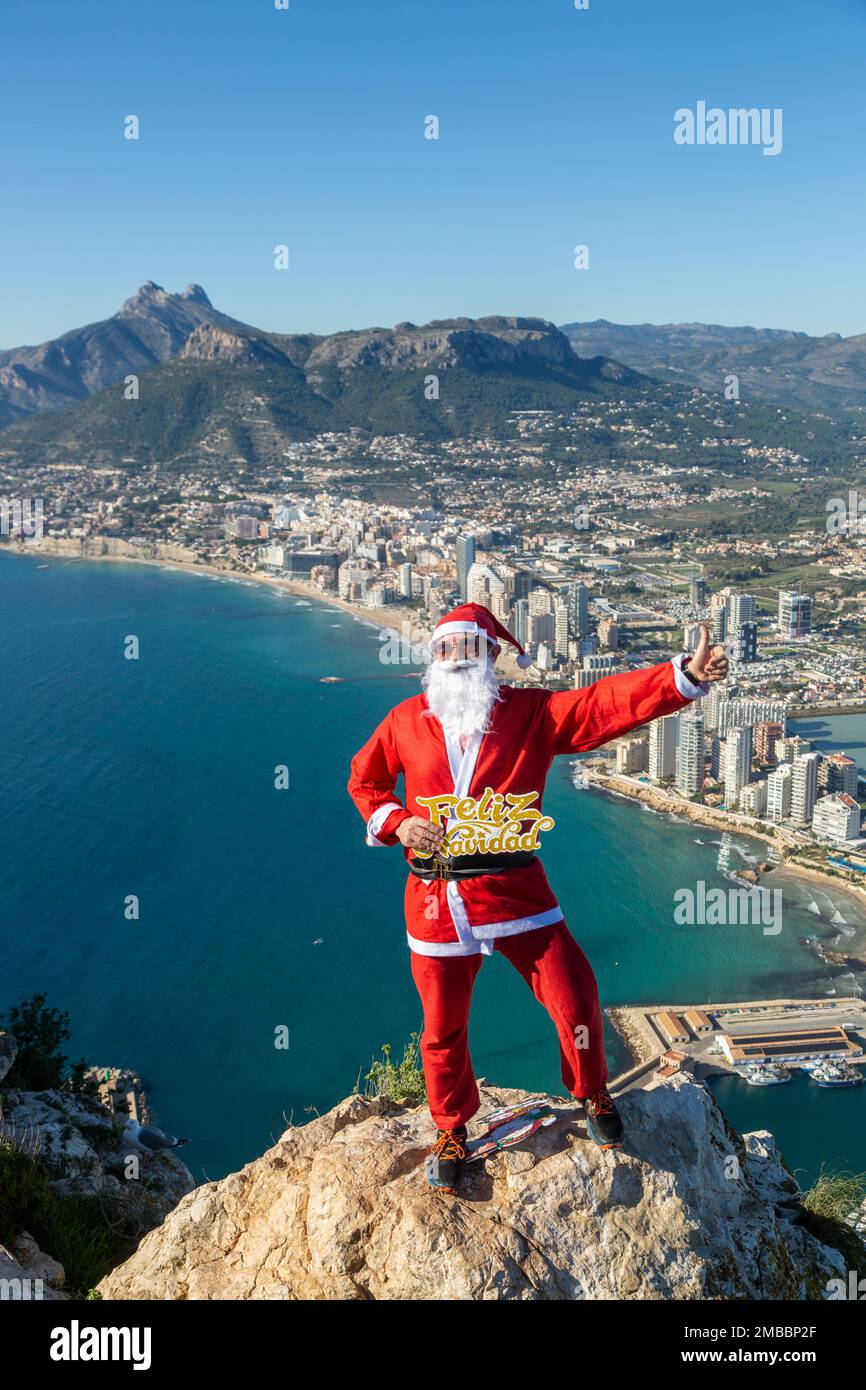 A man dressed as Santa Claus holding a sign saying feliz navidad standing on top of mountain. Stock Photo
