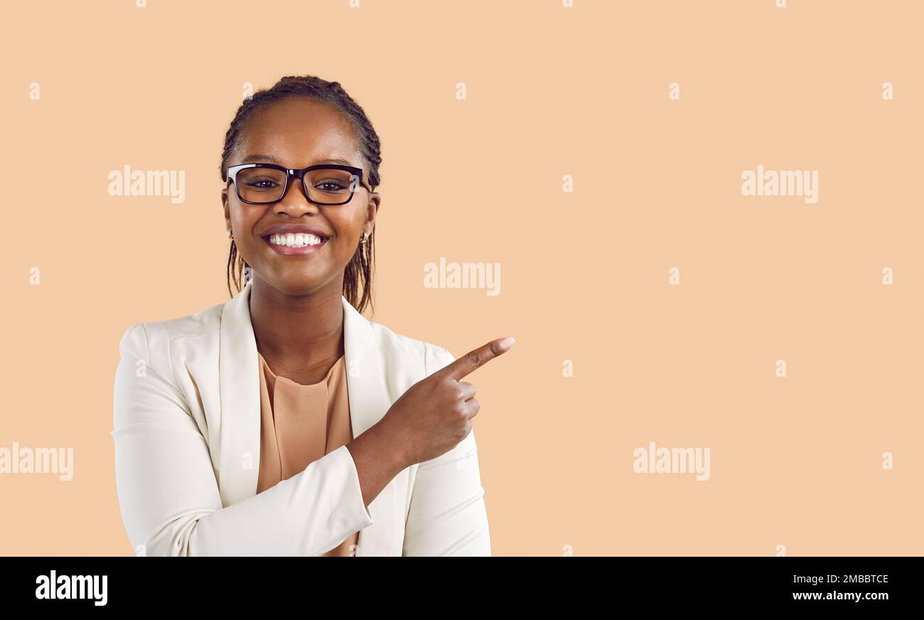 Smiling female client looks at you and points her index finger at copy space on beige background. Stock Photo