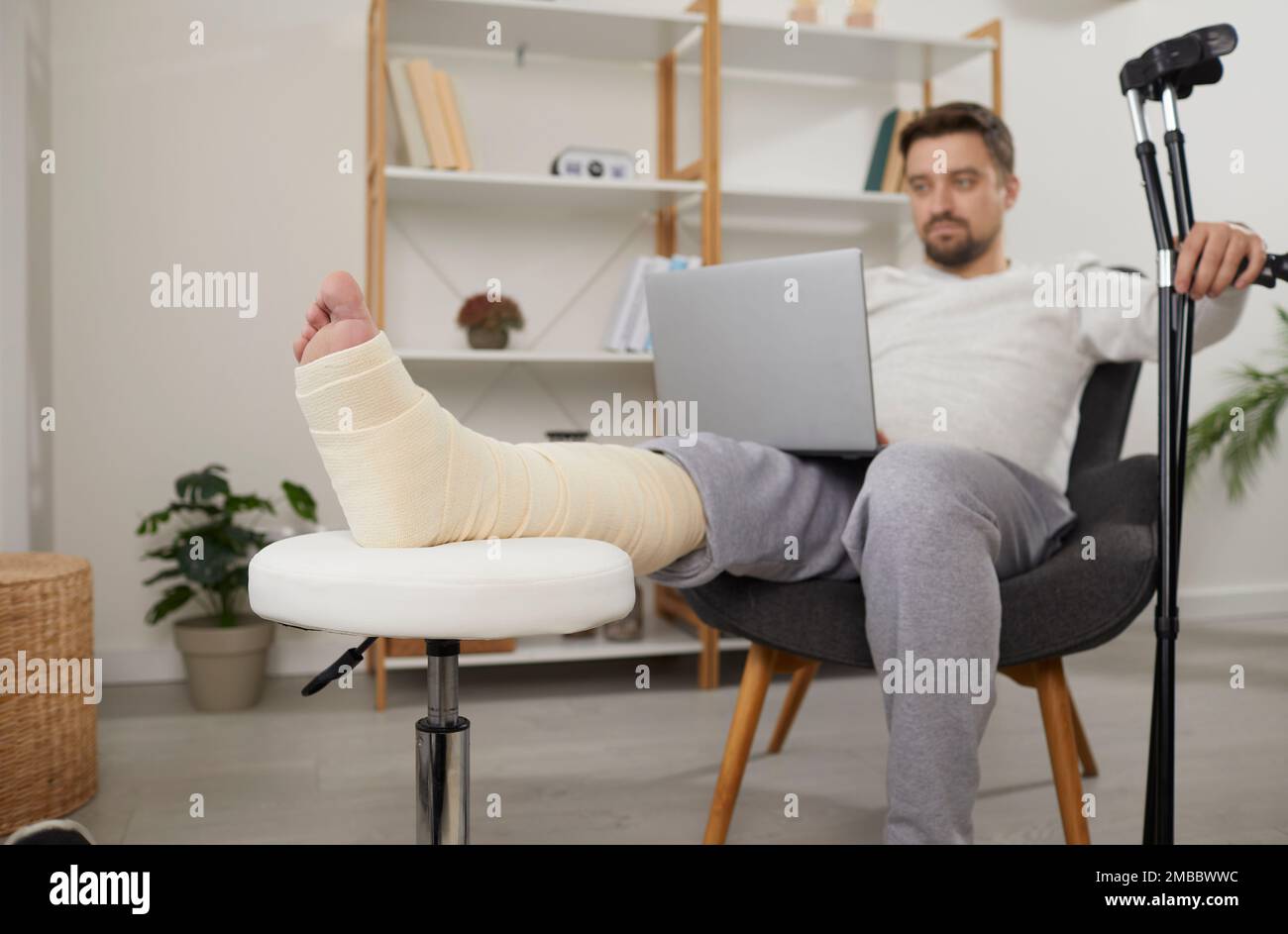 Man with broken leg sitting in armchair using laptop computer Stock Photo