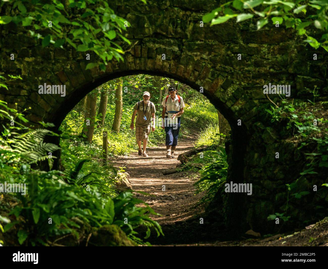 Two walkers approach small, single arch stone and brick bridge over old trackway in Ticknall Limeyards, Derbyshire, England, UK. Stock Photo