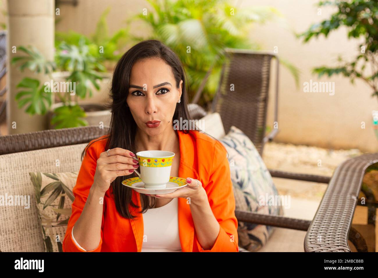 Goiania, Goiás, Brazil – January 10, 2023: A young woman, having tea, sitting in a chair with a calm expression, on the porch of her house. Half body Stock Photo