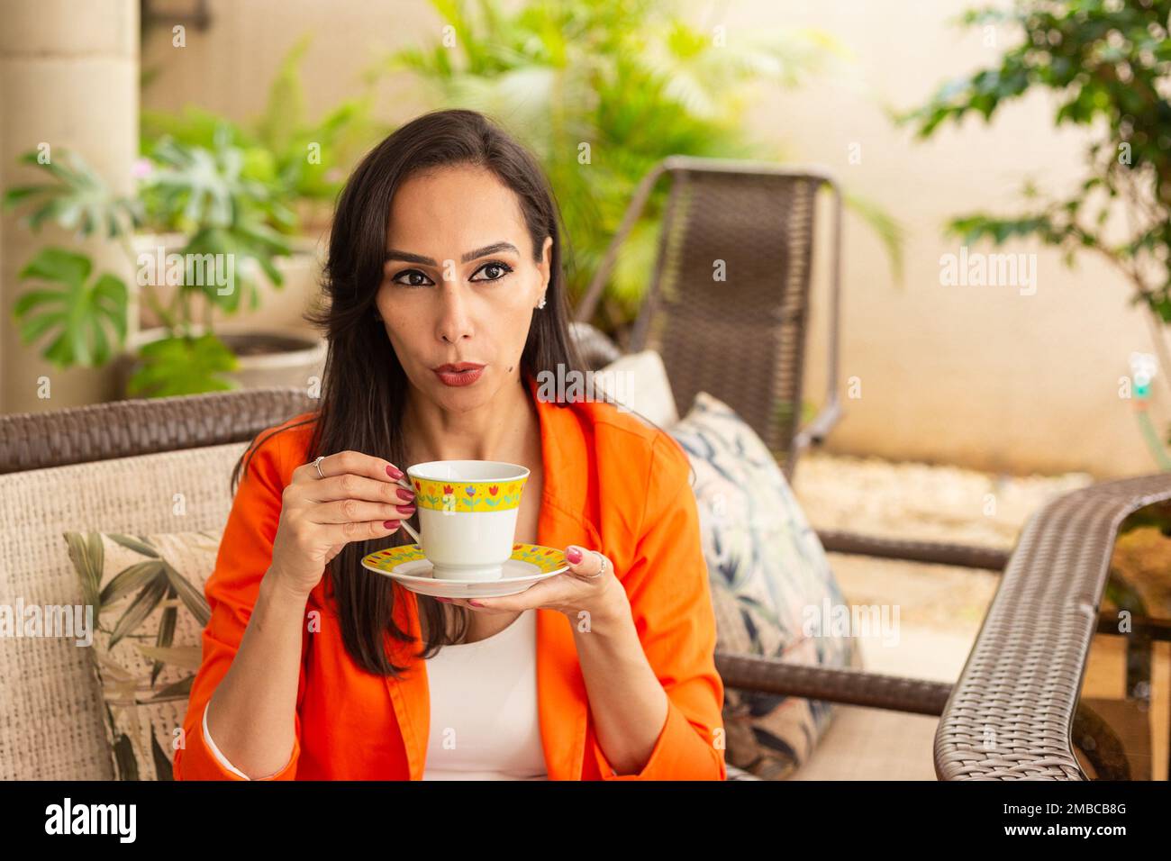 Goiania, Goiás, Brazil – January 10, 2023: A young woman, having tea, sitting in a chair with a calm expression, on the porch of her house. Stock Photo