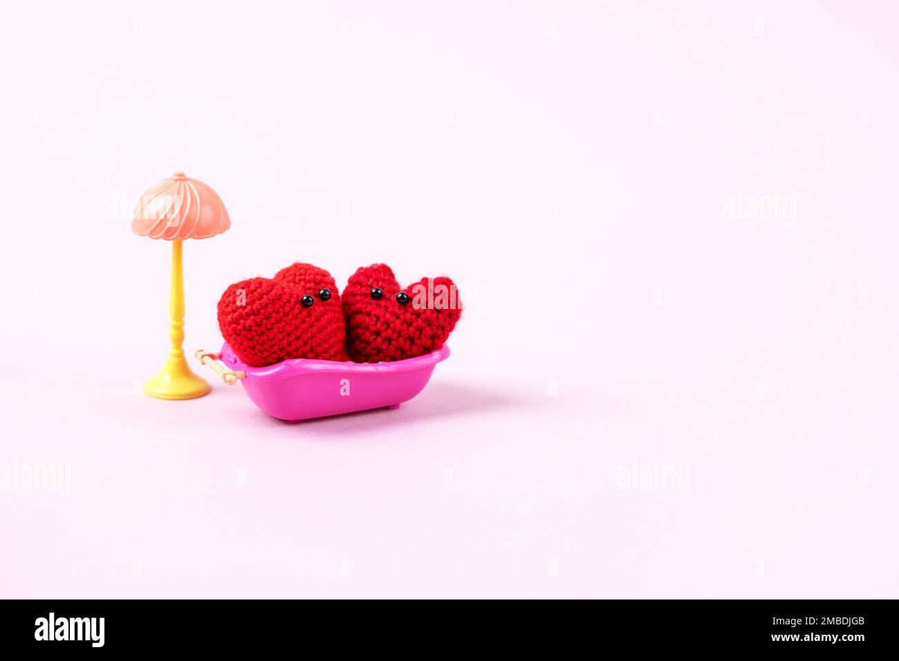 Concept of Valentine's Day. A family of two knitted red hearts in a pink toy bath and a toy lamp on a lilac background. Symbol of love, family, loyalt Stock Photo